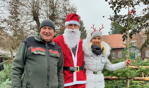 3 Personen vor einem Weihnachtbaumverkauf. die mittlere ist als Weihnachtsmann verkleidet